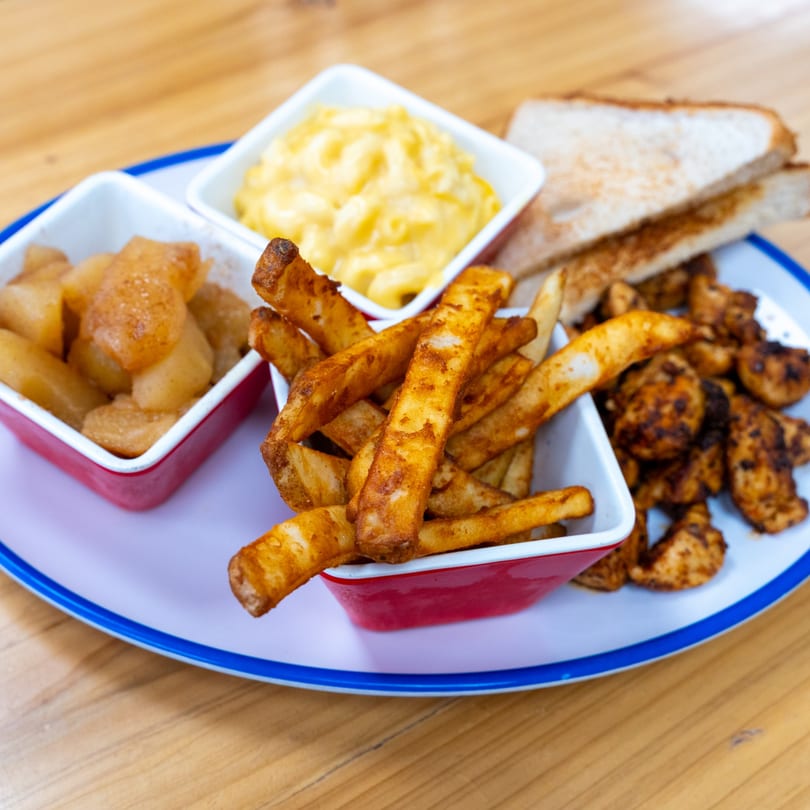 A plate of fries, mac and cheese, cinnamon apples, chicken bites, and toast on a wooden table.