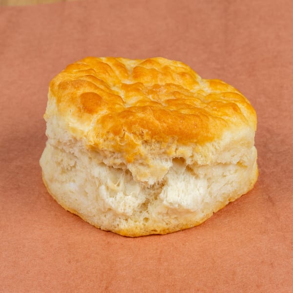 A golden-brown, fluffy biscuit sits on a rustic brown surface, ready to be enjoyed.