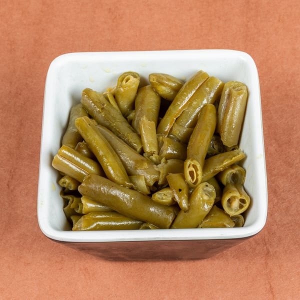 A white square bowl filled with cooked green beans, glistening with liquid, on a brown surface.