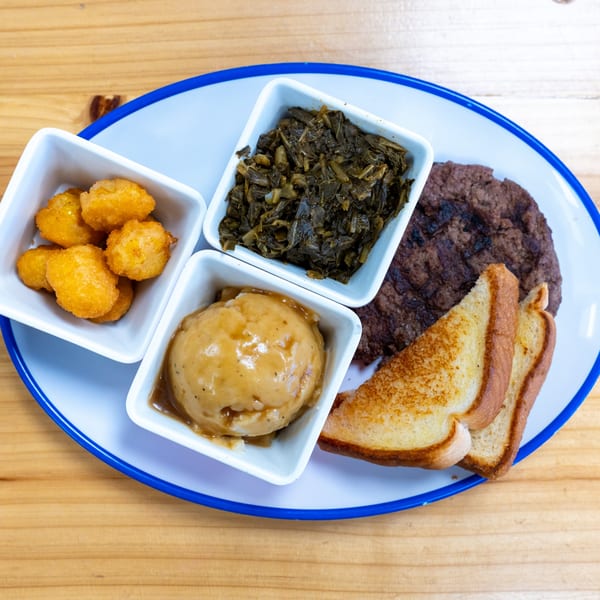 A Salisbury steak with mashed potatoes, collard greens, tater tots, and Texas toast on a plate.