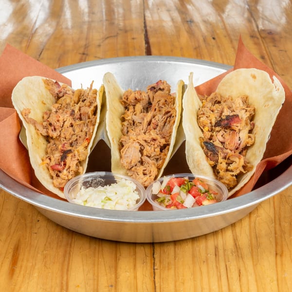 Three pulled pork tacos with coleslaw and pico de gallo in a metal tray.
