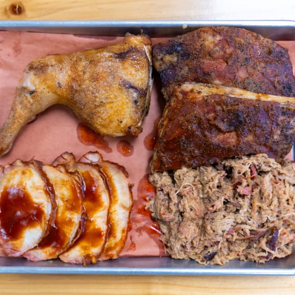 A metal tray filled with barbecue meats: chicken leg, ribs, sliced pork, and pulled pork.