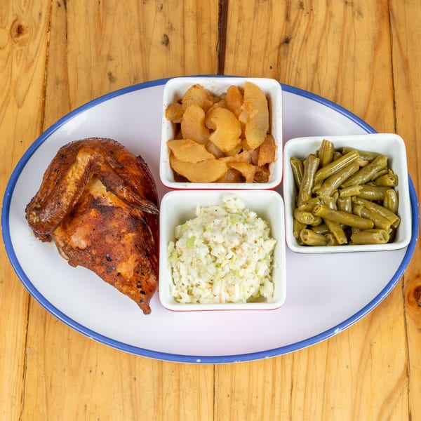 A plate with a roasted chicken quarter, coleslaw, green beans, and cinnamon apples on a wooden table.