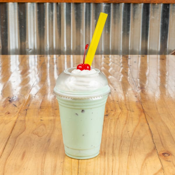 Mint green milkshake with whipped cream, a cherry, and a yellow straw on a wooden table.