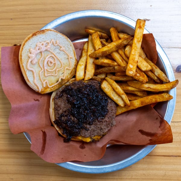 A deconstructed burger with caramelized onions and fries in a metal tray on a wooden table.