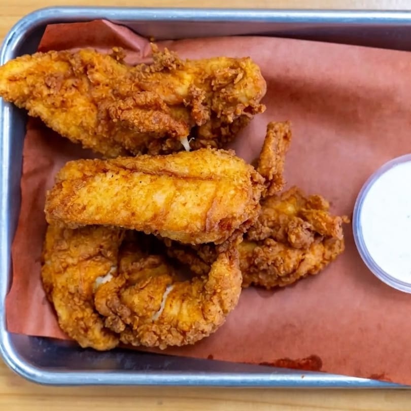 Crispy fried chicken tenders and a side of white dipping sauce in a metal tray.