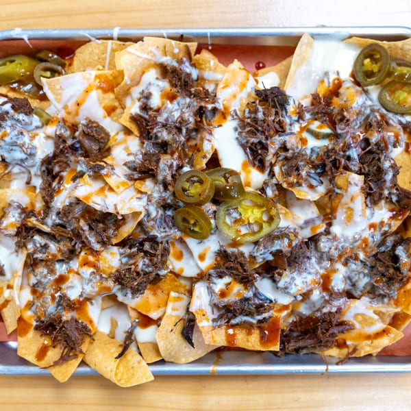 Close-up of loaded nachos with shredded beef, white sauce, barbecue drizzle, and jalapeños.