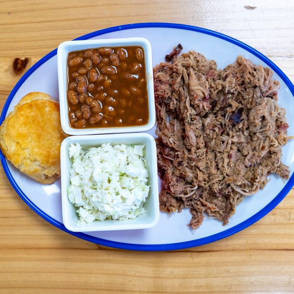 A plate of pulled pork, baked beans, coleslaw, and a biscuit on a wooden table.