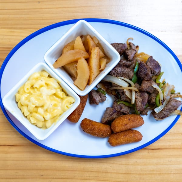 Steak with peppers and onions, mac and cheese, cinnamon apples, and hush puppies on a white plate.