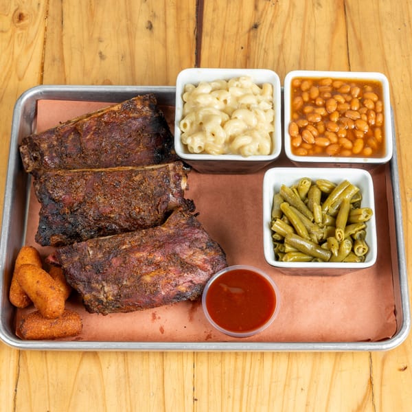 Smoked ribs, hush puppies, macaroni and cheese, baked beans, green beans, and BBQ sauce on a tray.