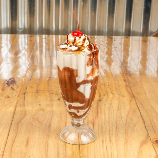 A tall glass of chocolate milkshake topped with whipped cream, chocolate drizzle, and a cherry.