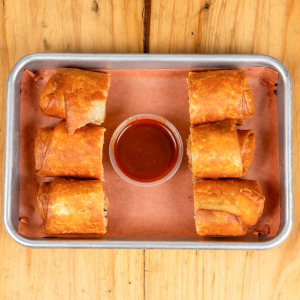 Six golden-brown egg rolls arranged in a metal tray with a small cup of dipping sauce in the center.